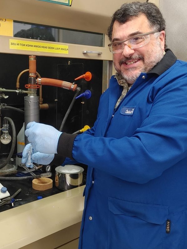 Dr. Mark Scialdone in a blue lab coat in front of lab glassware